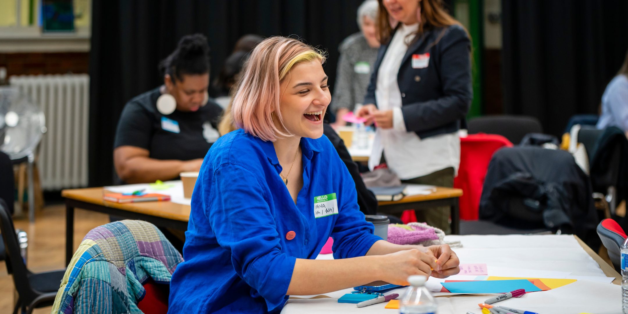 A person with pink hair that reaches their chin is wearing a bright blue shirt. They are smiling and leaning on a table covered with art supplies like post it notes and pens. There are people in the background - it looks like a fun, educational event